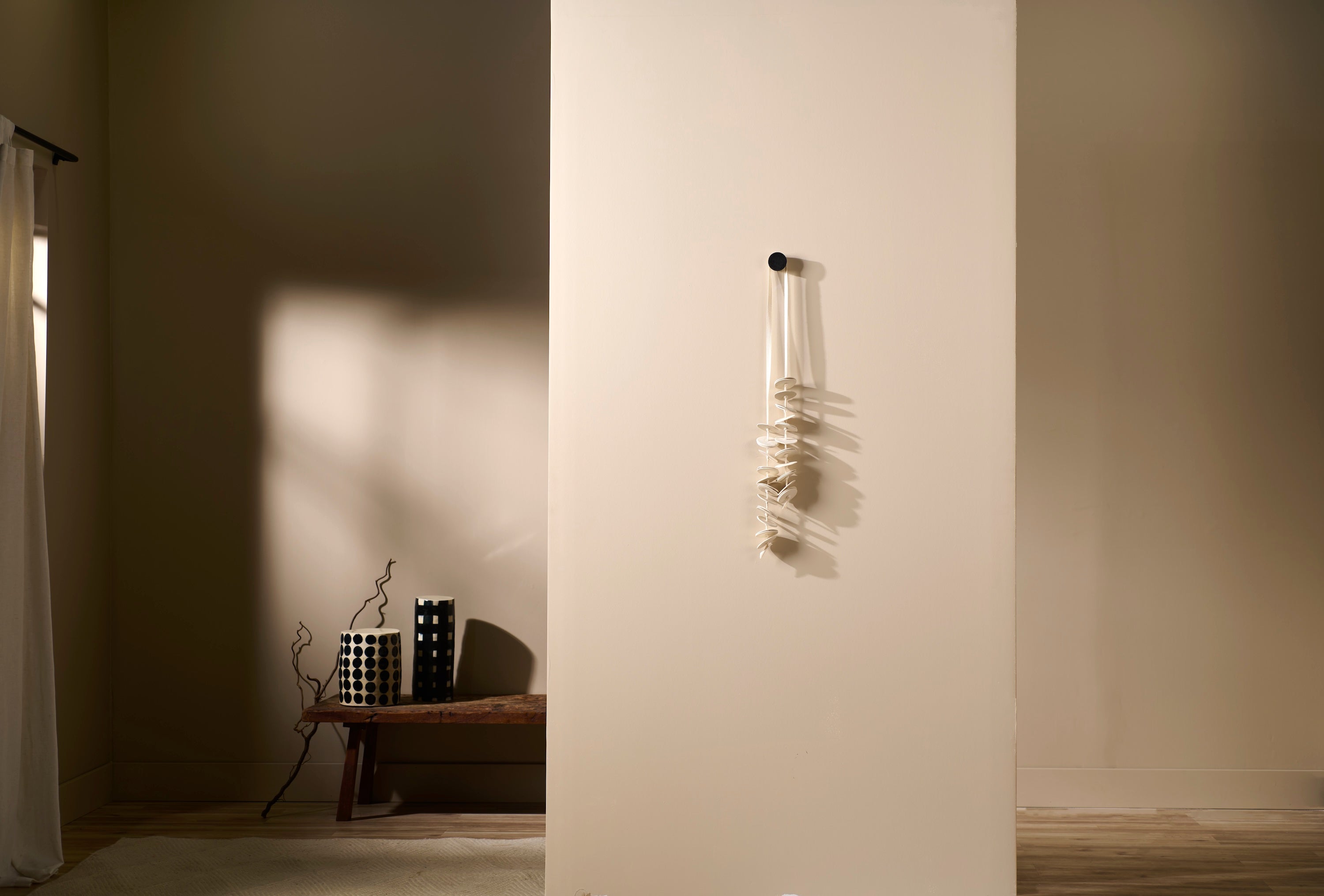 Vertical porcelain wall sculpture Cadence 2 by Lauren HB Studio installed on a soft beige wall. Handmade porcelain discs hang from a single knob, casting organic shadows that emphasize rhythm and movement.