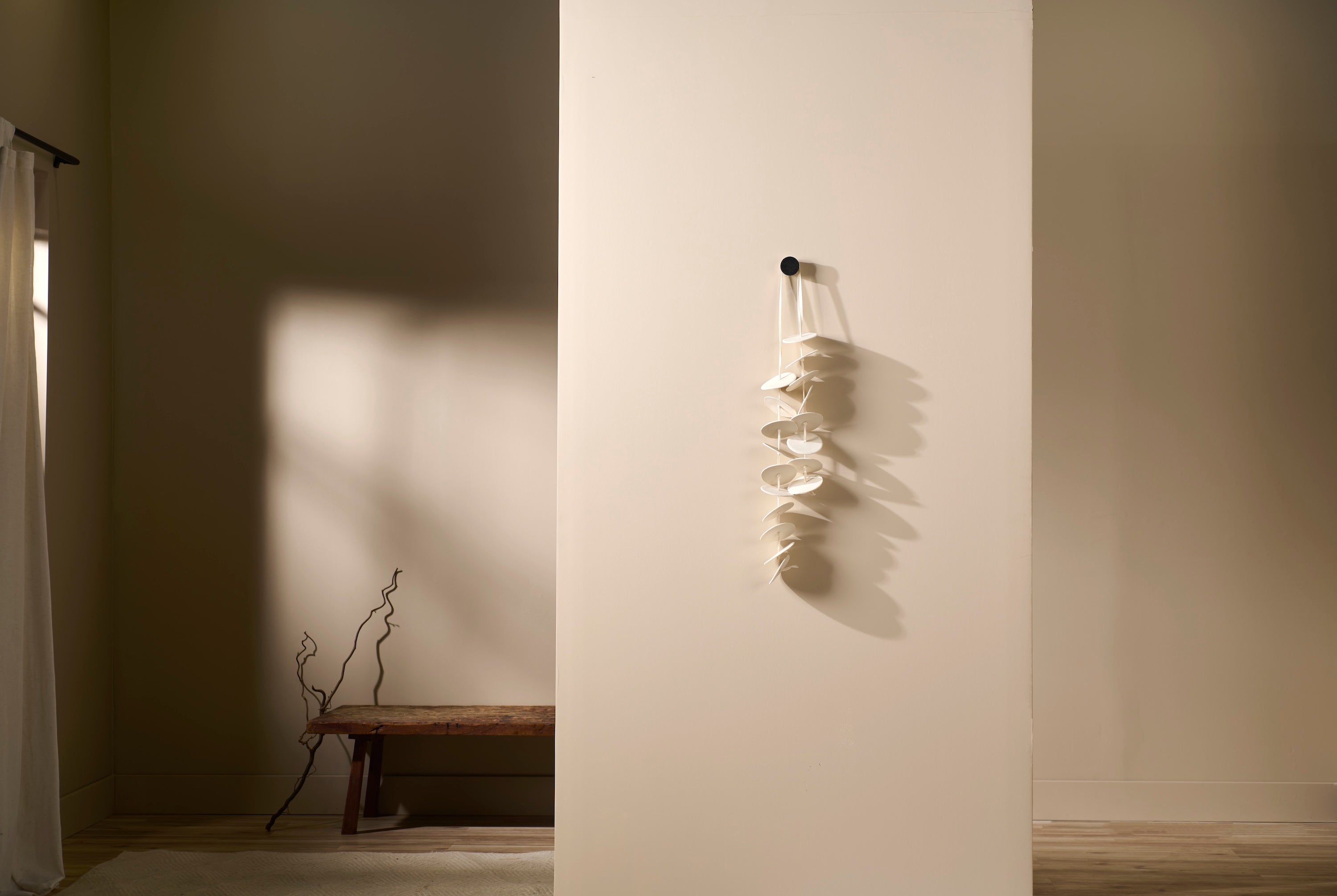 Vertical porcelain wall sculpture Cadence 3 by Lauren HB Studio displayed on a neutral wall. Handmade discs suspended from a single knob create rhythmic shadows.