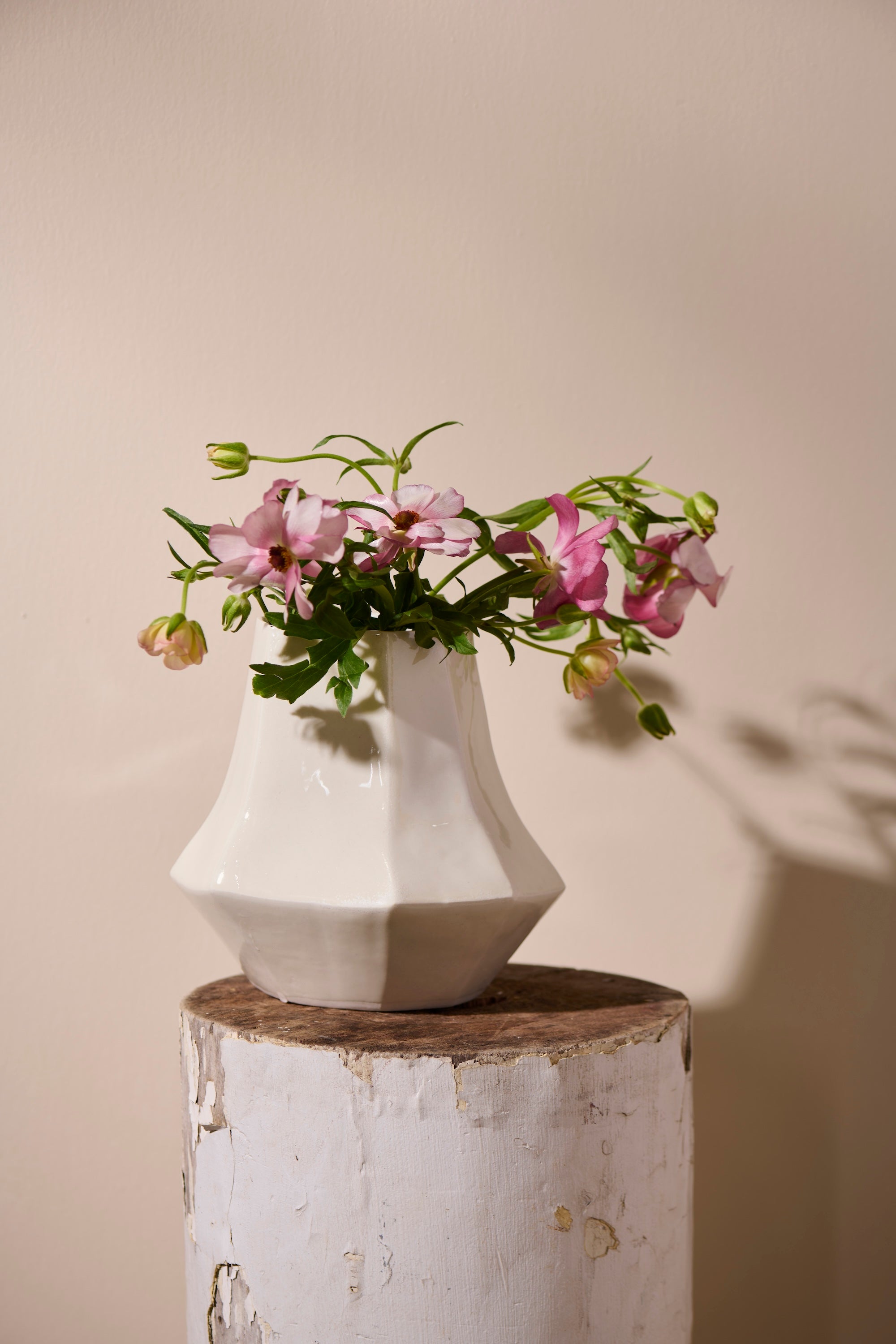 Lantern Vase by Lauren HB Studio — handmade porcelain vase with geometric planes finished in Frost White glaze, displayed with delicate pink flowers atop a tall cream pedestal against a neutral wall. The composition highlights its faceted form and luminous glaze, evoking the shape of a modern lantern.