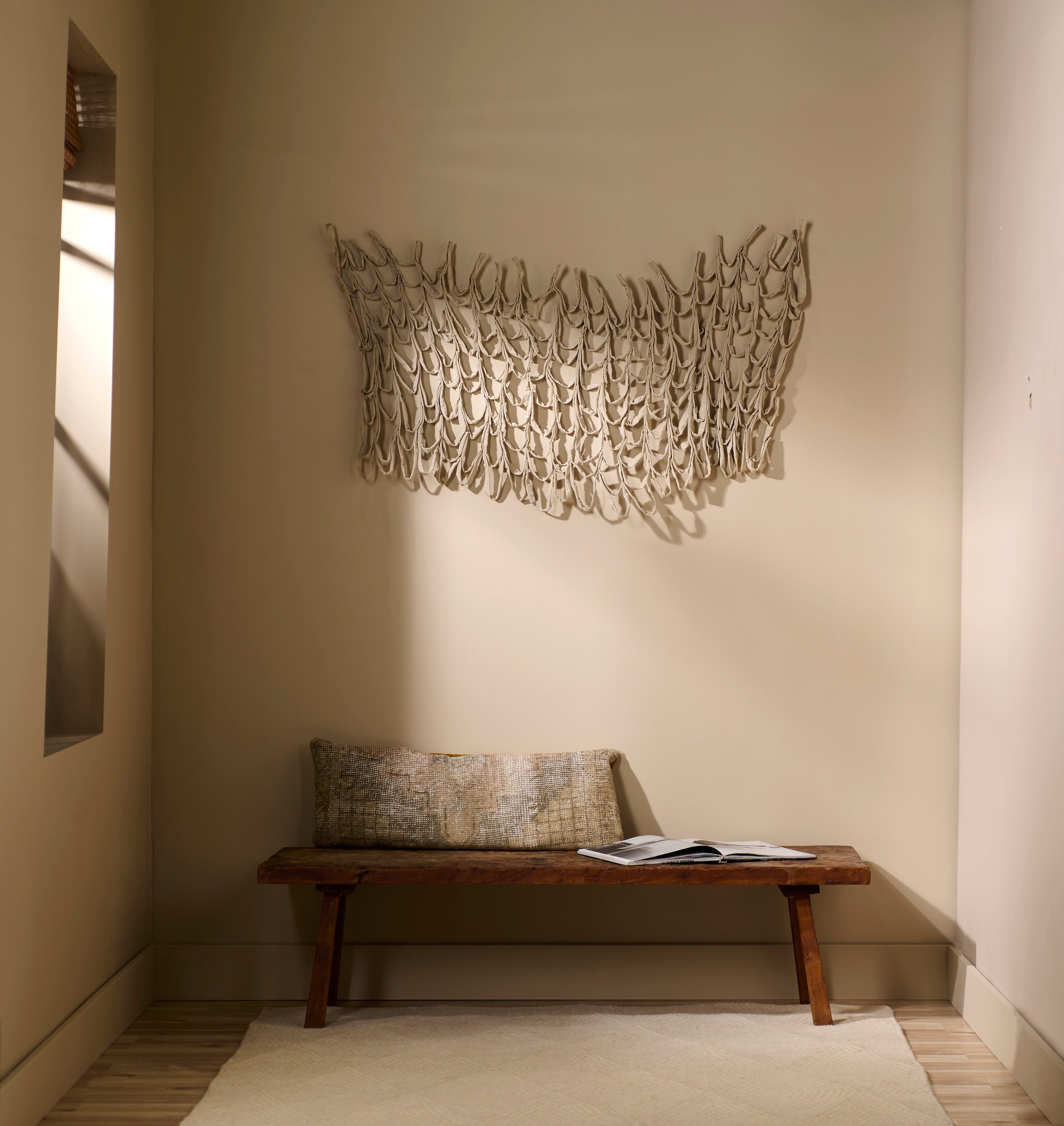 Murmur 2 fabric wall sculpture by Lauren HB Studio — a layered textile installation in soft tawny hues, mounted above a wooden bench against a neutral wall. The handsewn forms cascade in gentle loops, creating movement and texture reminiscent of wind or tide.