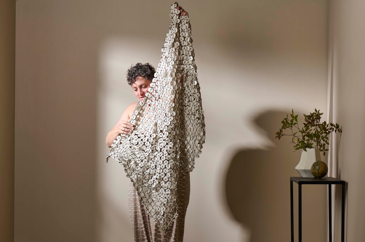 The artist Lauren Herzak-Bauman holding Woven wall sculpture made of porcelain discs, standing in a room with a wood table and vessels with. neutral wall.